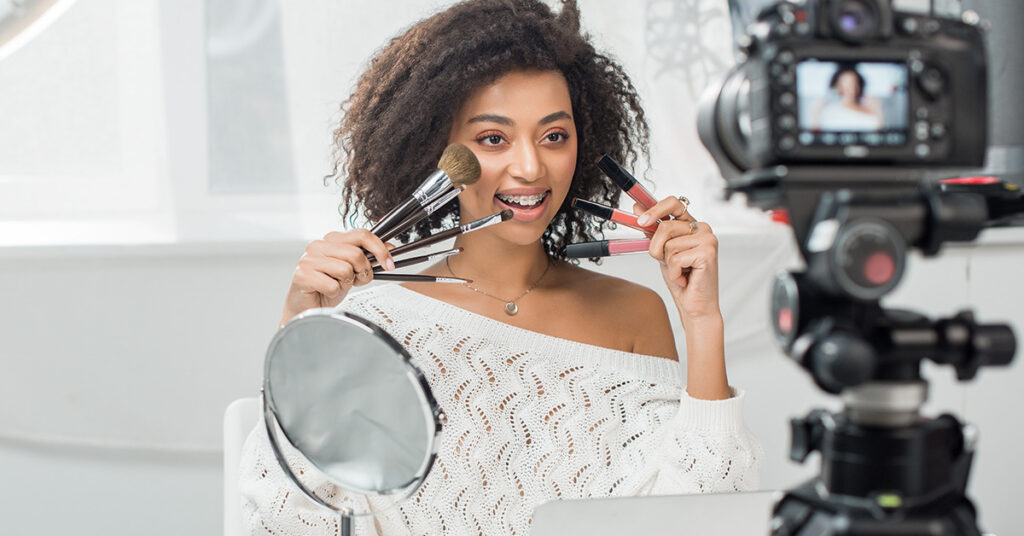 girl with curly brown hair lifting makeup brushes to her face whilst filing a video